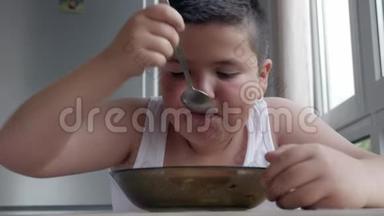 可爱的小胖男孩在厨房里吃饭，吃一大勺汤，儿童肥胖和暴食的概念